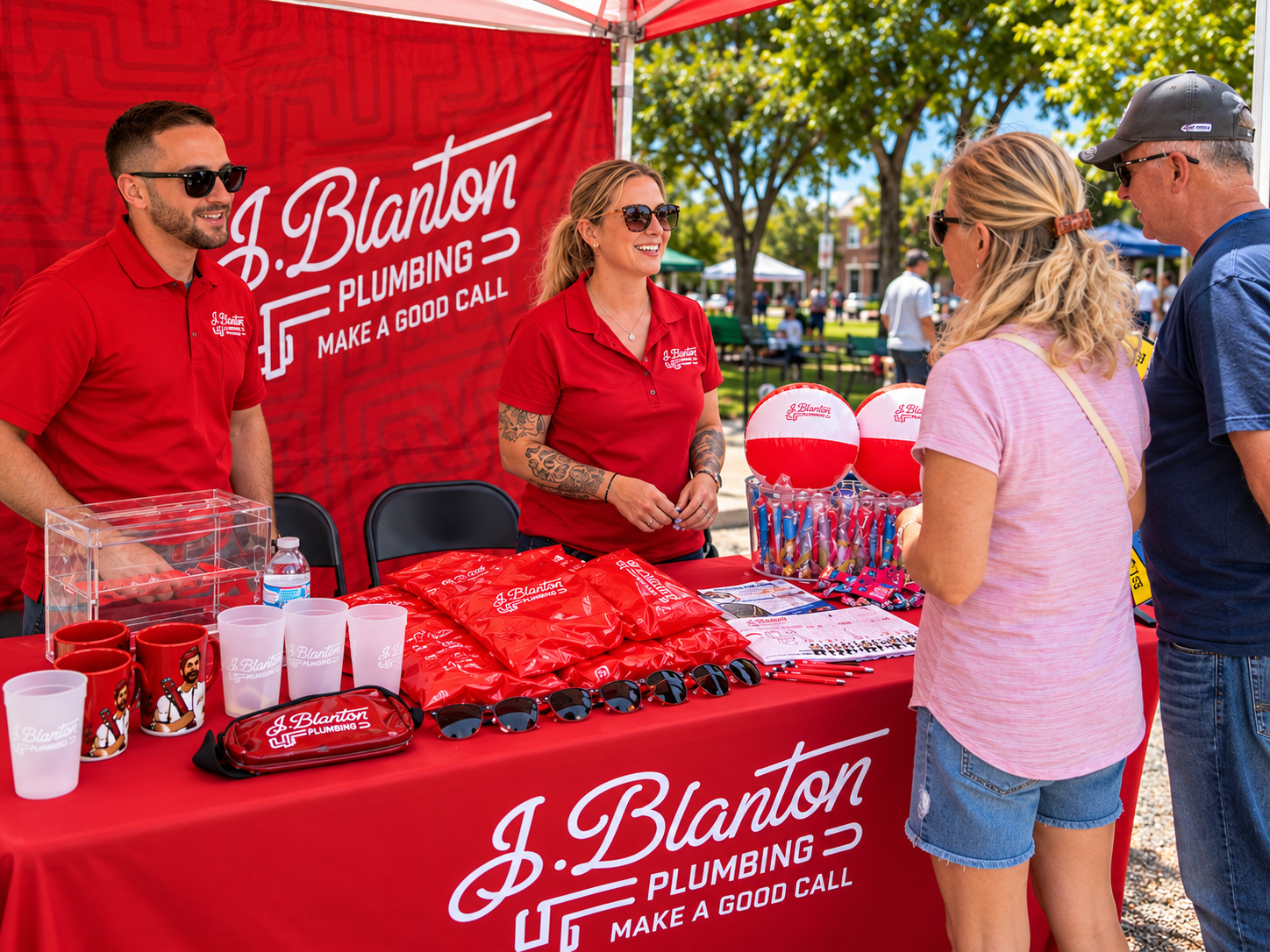 J. Blanton Plumbing booth at a community fair with branded table, mugs, sunglasses, and team members speaking with visitors